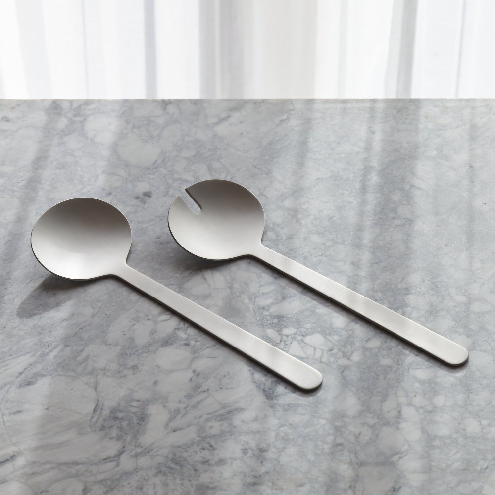 Two stainless steel serving utensils, one spoon and one fork spoon with a small notch at the tip, resting on a gray marble surface with a soft-focus white curtain background
