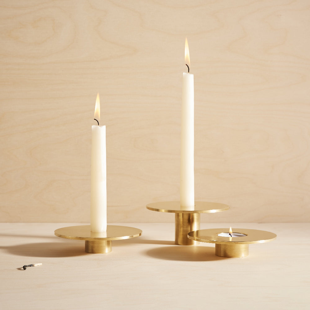Three gold brass candle holders of varying heights and widths, each holding a lit white taper candle, arranged on a light wood surface against a matching light wood background.