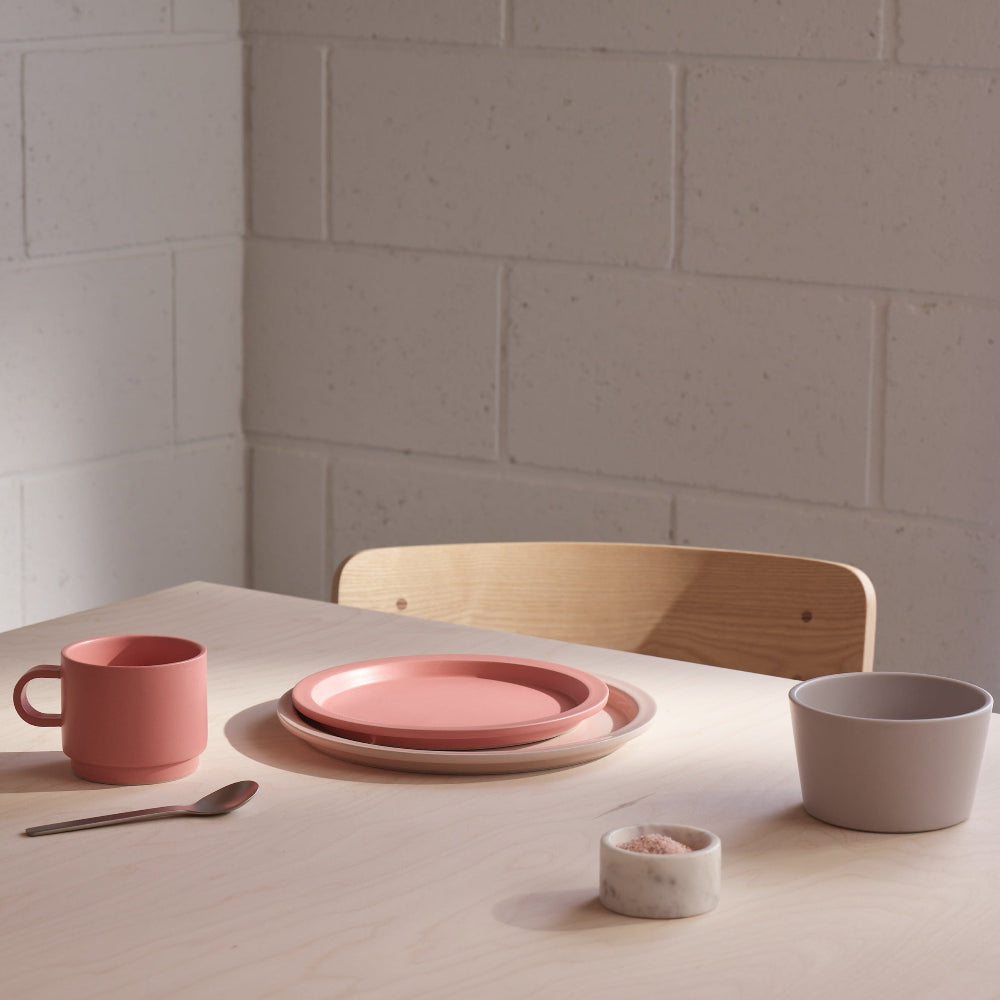 A styled dining table setting featuring a dusty pink ceramic mug, matching pink plates, a light wood chair, and a neutral gray bowl on a light wood table against a white brick wall.