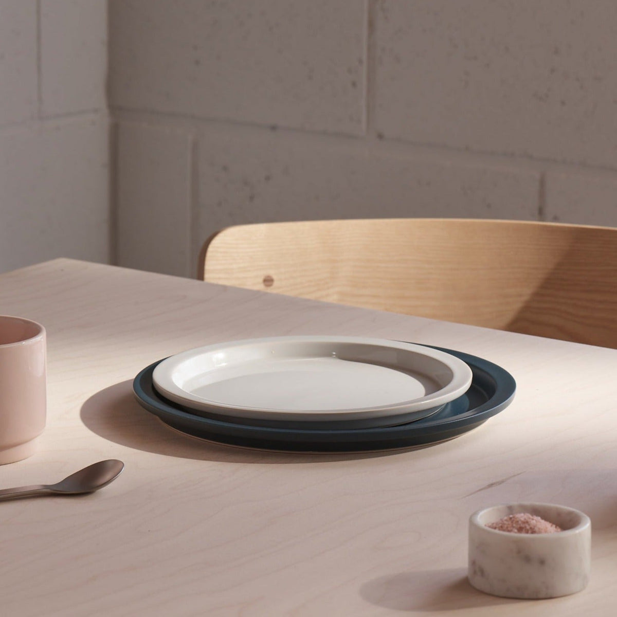 A stack of two ceramic plates, one white and one dark teal, set on a light wood table in a minimalist dining setting with a wooden chair, a pink mug, a spoon, and a small salt dish.