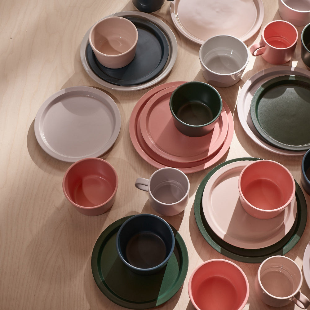 A styled flat-lay collection of matte ceramic tableware in muted pastel and earthy tones, including plates, bowls, cups, and mugs in pink, green, blue, gray, and beige, arranged on a light wood surface.