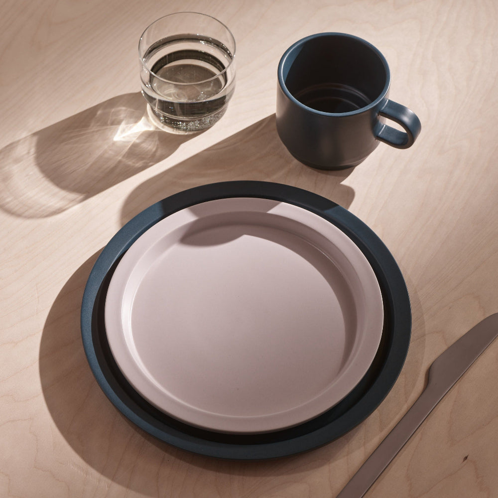 A minimalist ceramic table setting on a light wood surface, featuring a round plate with a dark blue rim and a pale pink center, a matching mug, a clear glass of water, and a knife.
