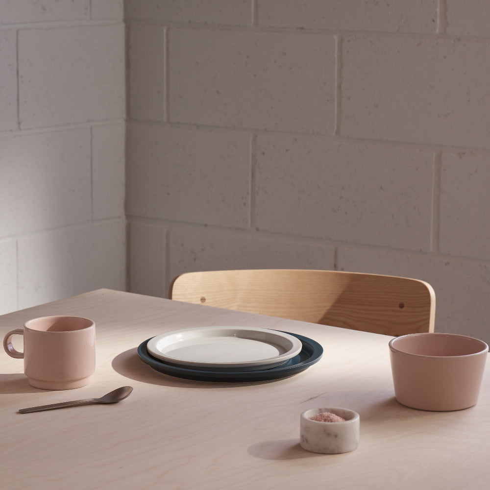 A styled dining table scene with a stack of ceramic plates, a pink mug, a pink bowl, a small pink cup, a spoon, and a marble salt dish against a light brick wall background.