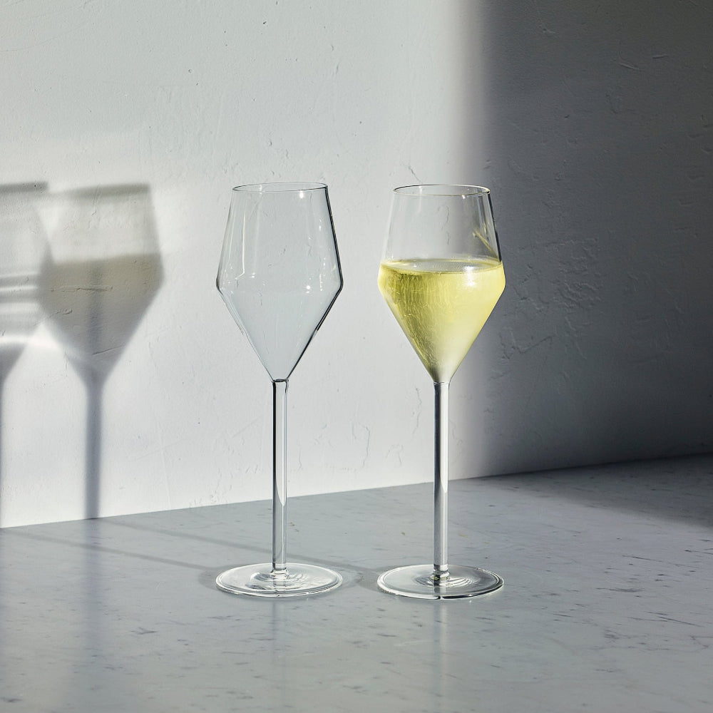 Two clear champagne glasses on a light gray marble surface against a white wall, with one glass empty and the other partially filled with champagne Strong sunlight casts shadows on the wall and surface.