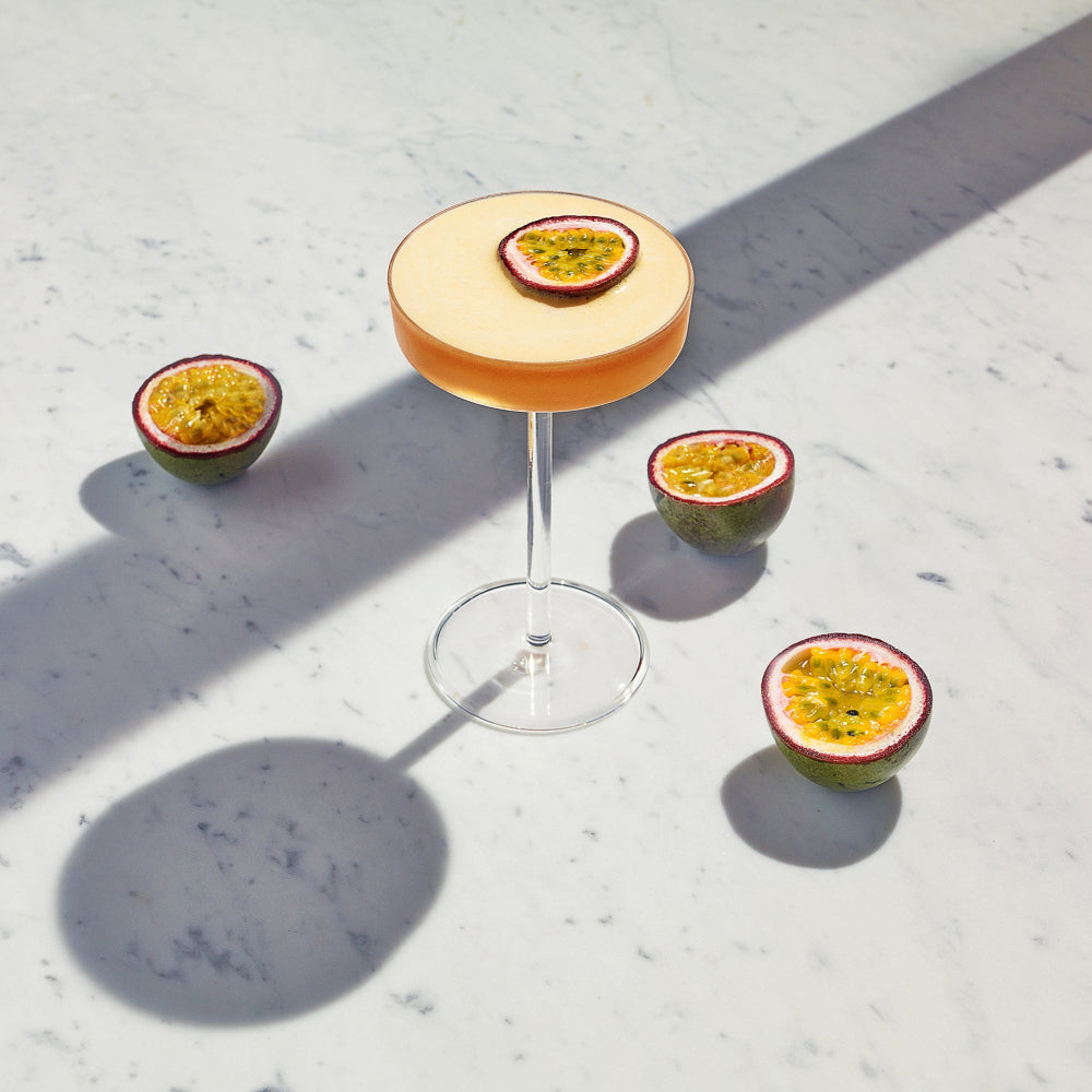 A stemmed cocktail glass filled with an orange-amber drink and topped with a sliced passion fruit garnish, arranged on a white marble surface with three additional passion fruit halves around it in bright sunlight.