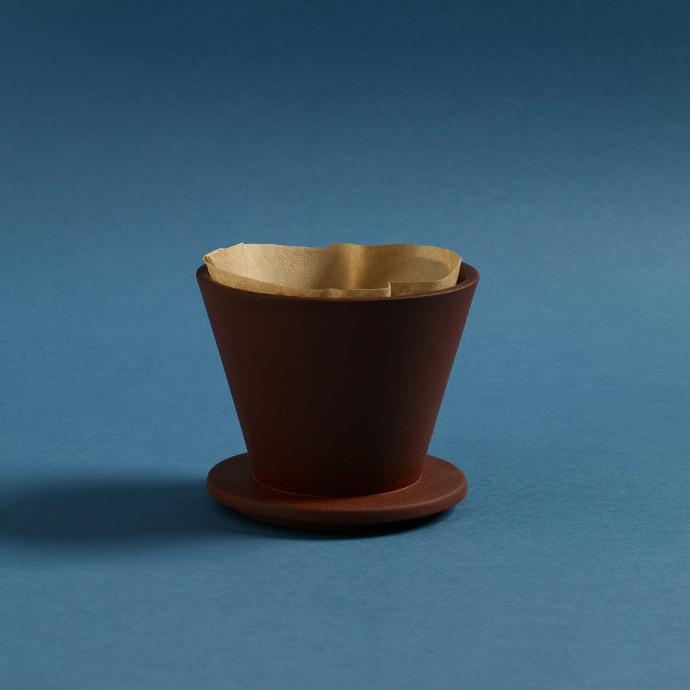 Terracotta COLOMBO ceramic coffee dripper by Aaron Probyn with a paper filter against a blue background