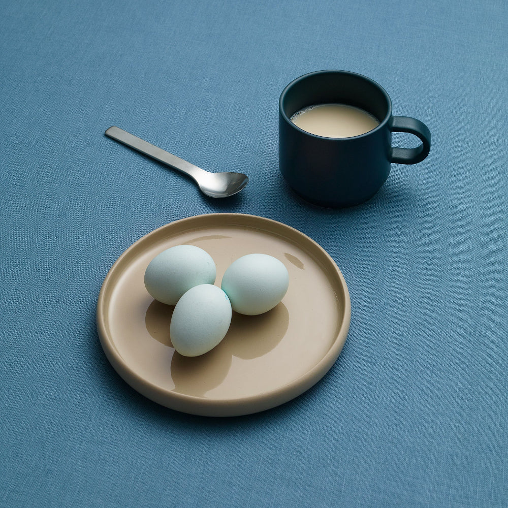 Three pale eggs on a beige plate next to a dark blue mug filled with a light beverage and a metal spoon, all arranged on a textured blue surface.