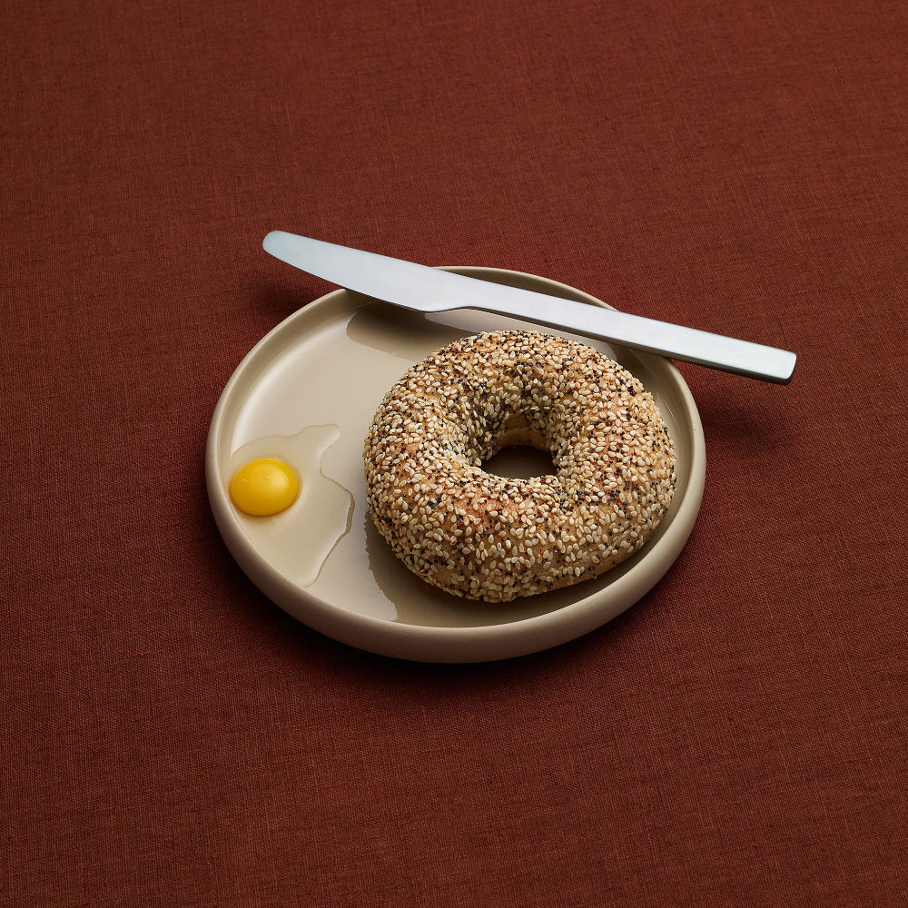 A sesame seed bagel on a small beige plate with a silver knife, with an uncooked egg on a textured dark brown background.