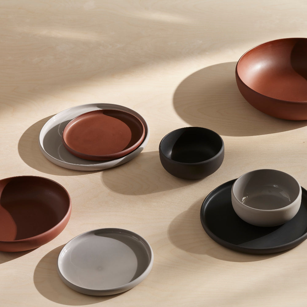 A set of matte ceramic tableware in earthy tones, including bowls, plates, and a small cup, arranged on a light wood surface with soft shadows.