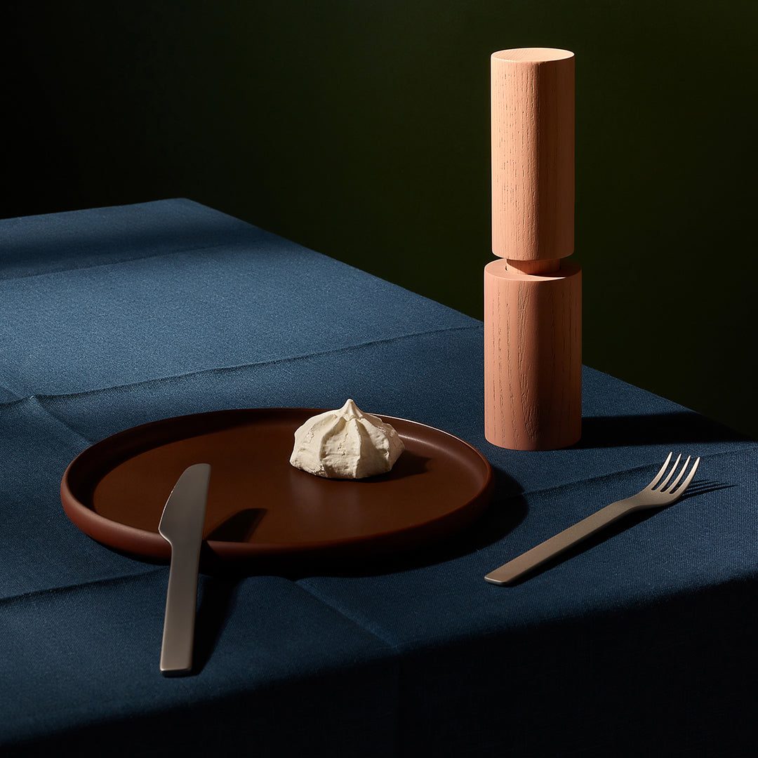 Ceramic plate with a small cake, knife, and fork on a dark blue surface with wooden cylindrical objects.
