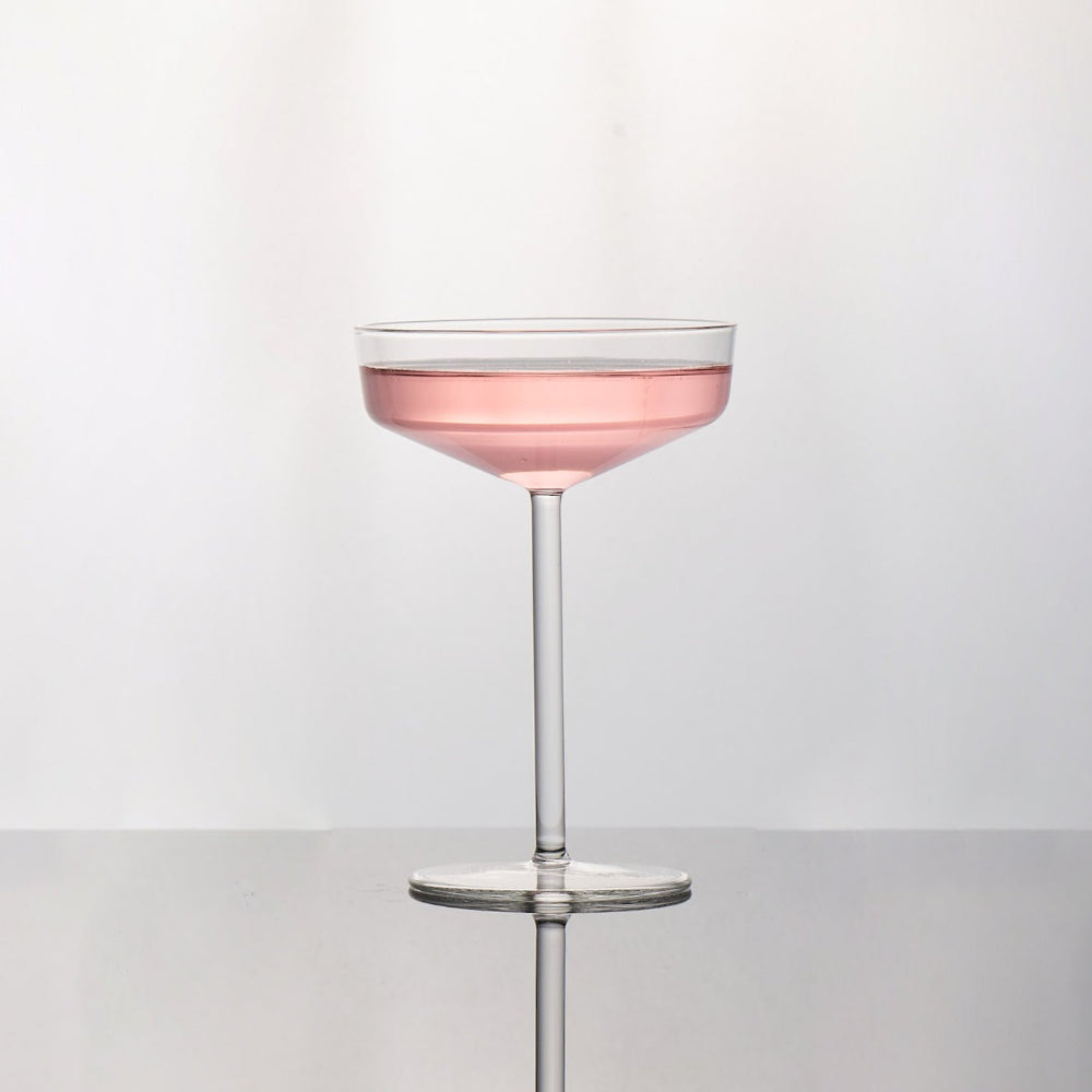 A clear stemmed coupe glass filled with pink liquid, centered on a light gray reflective surface against a white background.