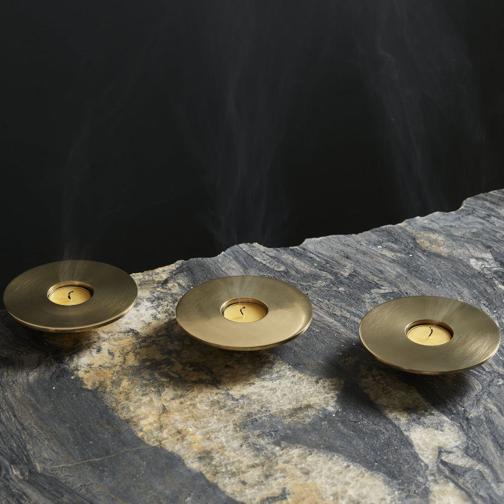 Three round brass candle holders with lit tealights, shown on a dark stone surface with smoke rising against a black background.