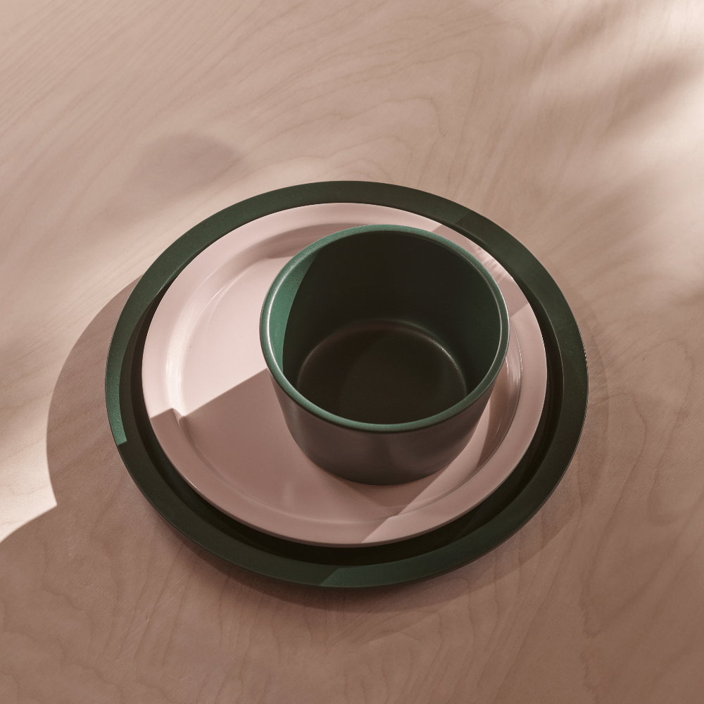 A minimalist ceramic table setting featuring a dark green cup nested inside a light pink plate, which is in turn nested inside a dark green charger plate, all arranged on a light wood surface with soft leaf shadows.