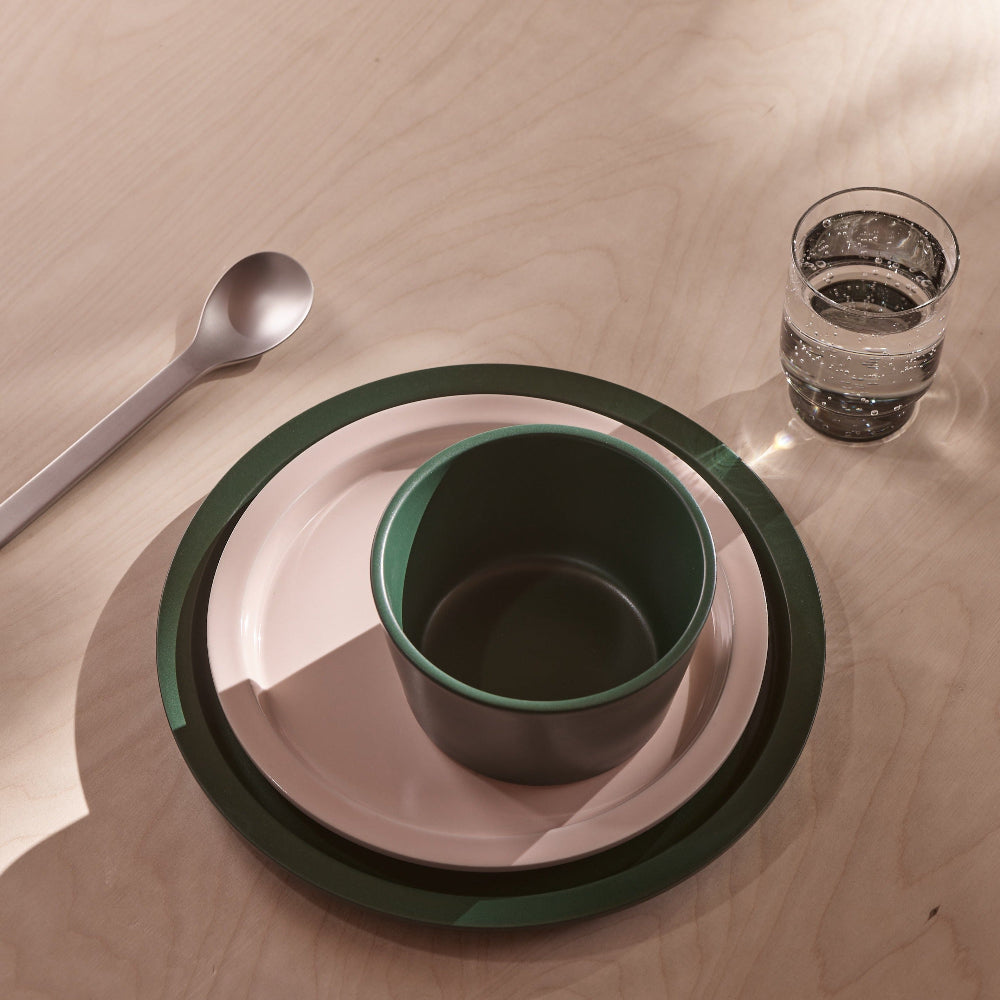 A place setting on a light wood tabletop featuring a stack of plates in dark green and pale pink, a matching dark green cup, a silver spoon, and a clear glass of water.