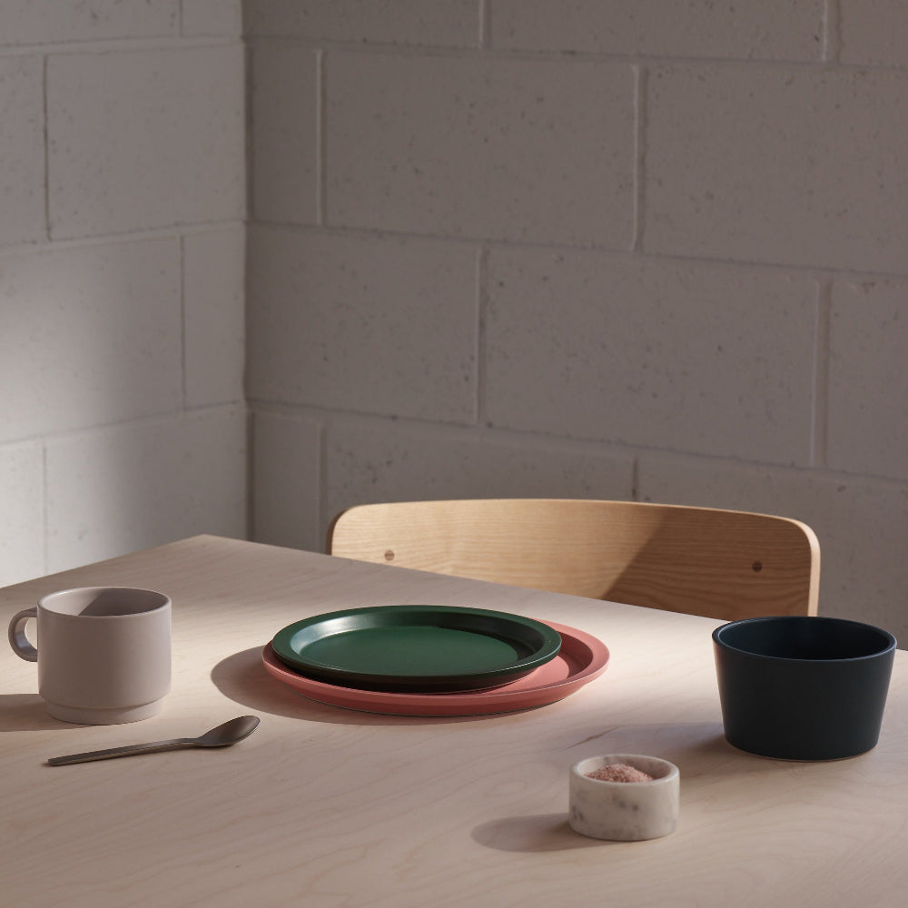 A minimalist dining table setting in a warm, sunlit room, featuring a stack of ceramic plates in dark green and terracotta pink, a light gray mug, a dark navy bowl, a small marble salt dish, and a wooden chair against a light gray brick wall.