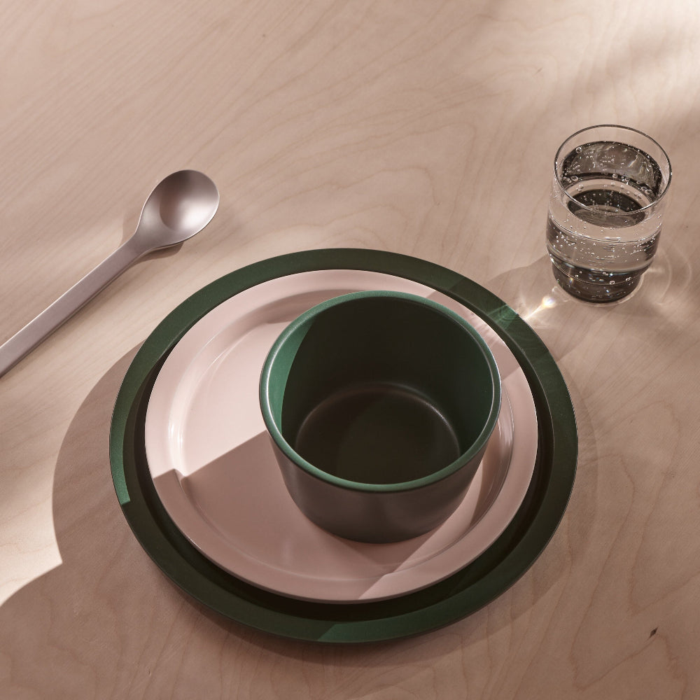 A place setting on a light wood tabletop featuring a dark green bowl nested inside a pale pink plate with a dark green rim, alongside a matching spoon and a clear drinking glass.