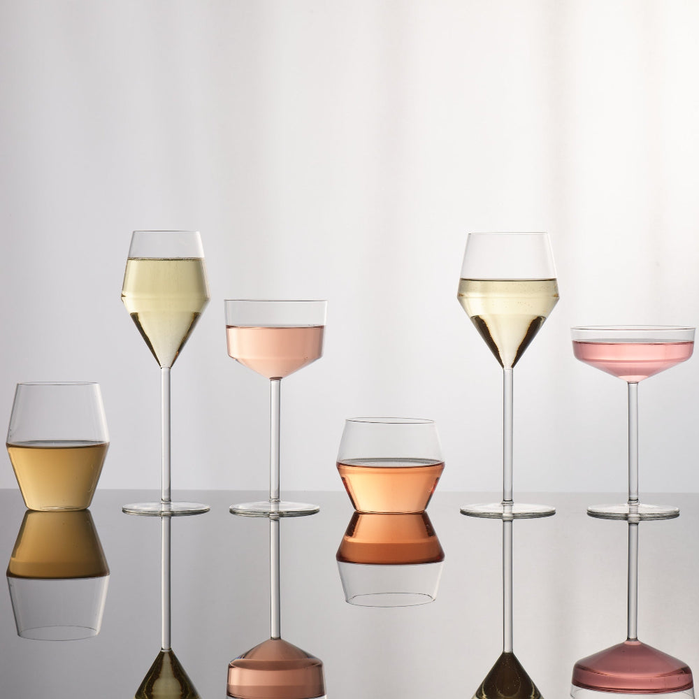 Six clear wine and cocktail glasses arranged on a reflective surface against a soft white background, each partially filled with different colored liquids: white wine, pale pink, amber, peach, pale pink, and white wine.