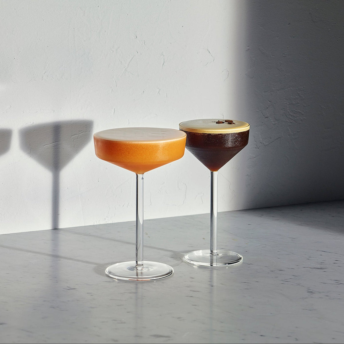 Two stemmed cocktail glasses filled with layered drinks, one orange-amber and one dark brown, set on a gray marble surface against a white wall with strong sunlight and shadows.