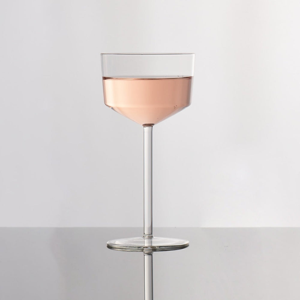 A clear stemmed cocktail coupe glass filled with pale pink liquid, centered on a reflective light gray surface against a white background.