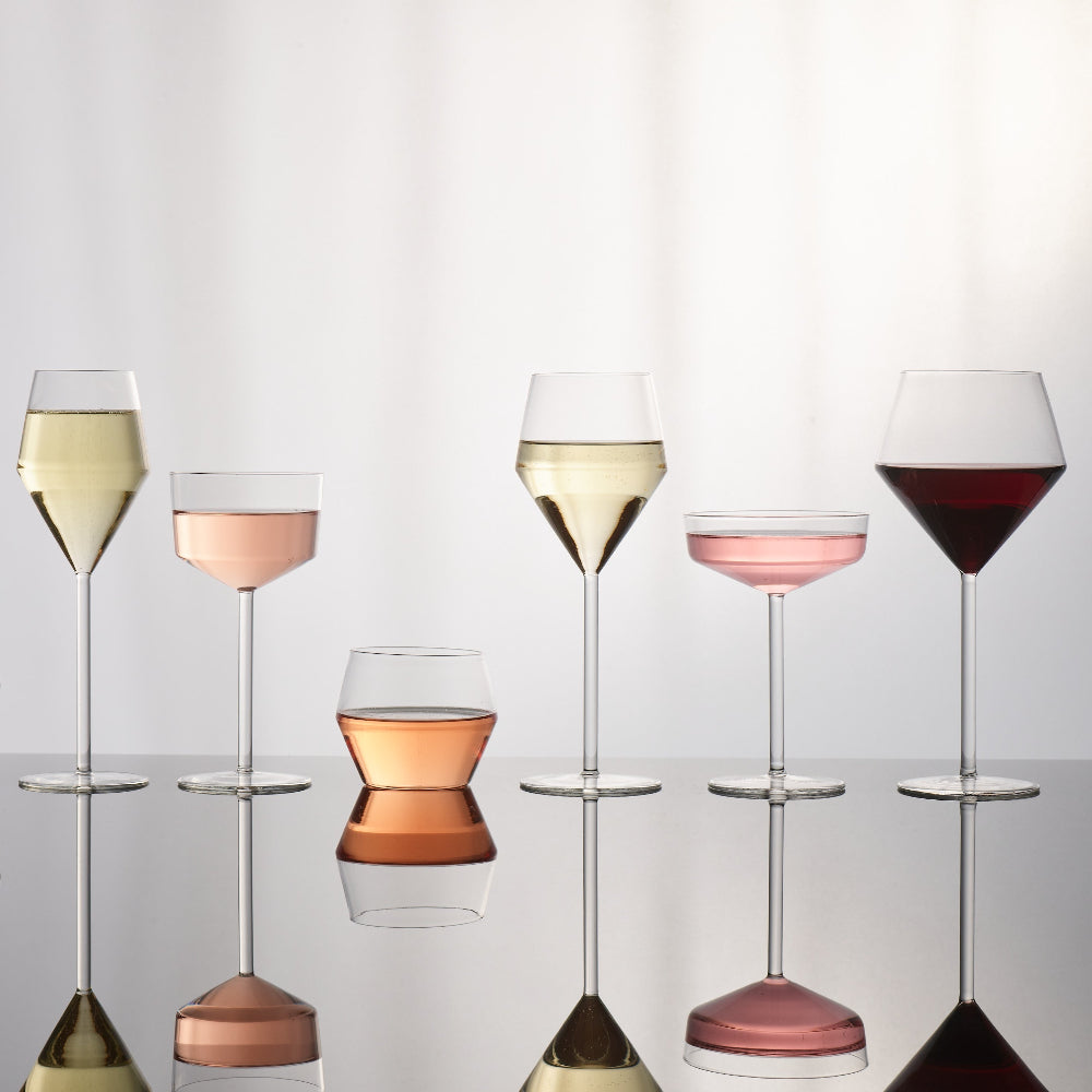 Six clear wine glasses in different shapes and fill levels, shown on a reflective surface against a soft white background. Some glasses are empty, while others are filled with white wine, rosé wine, and red wine, creating pink, peach, and deep red reflections.