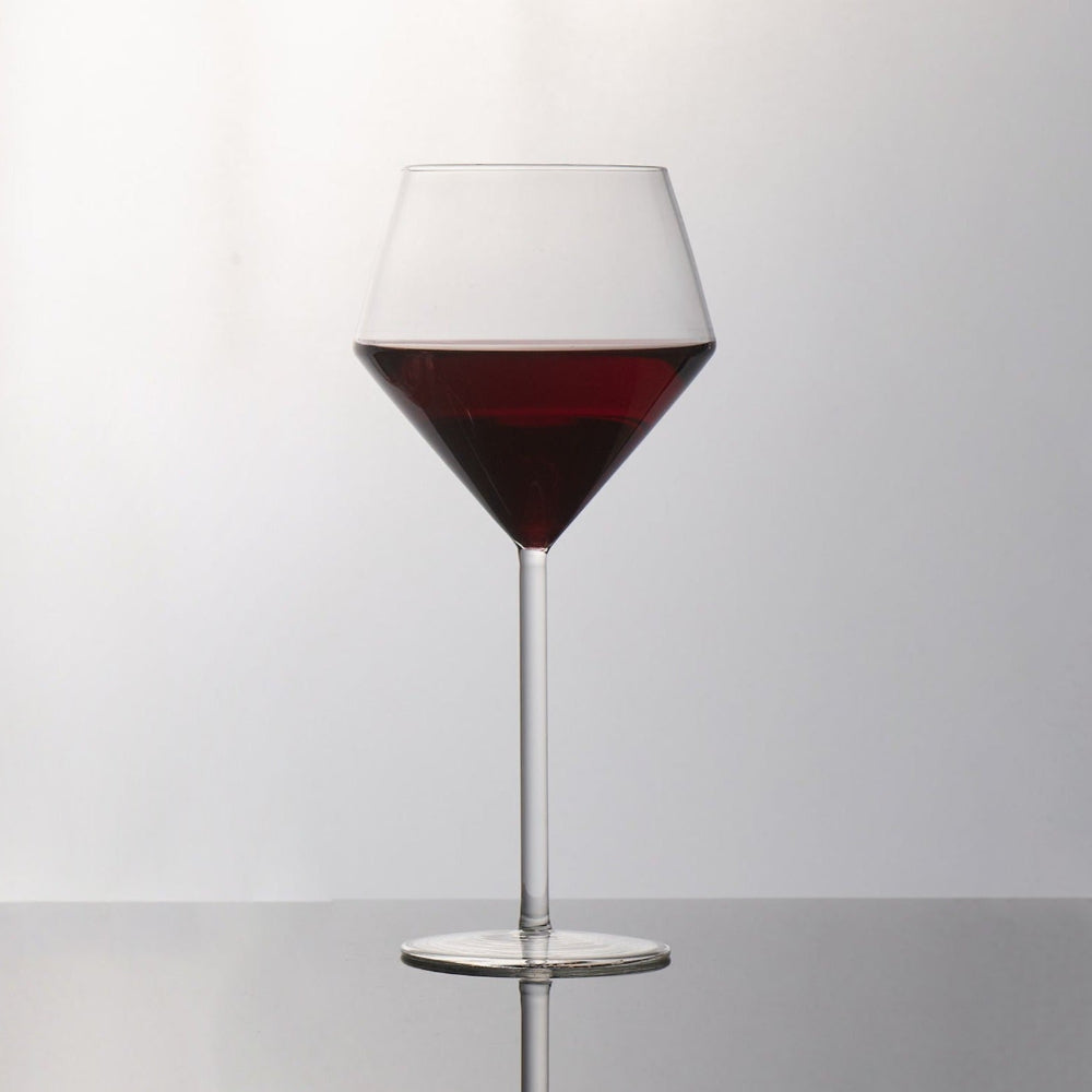 A clear stemmed wine glass with a wide bowl, partially filled with red wine, standing on a reflective surface against a light gray background.