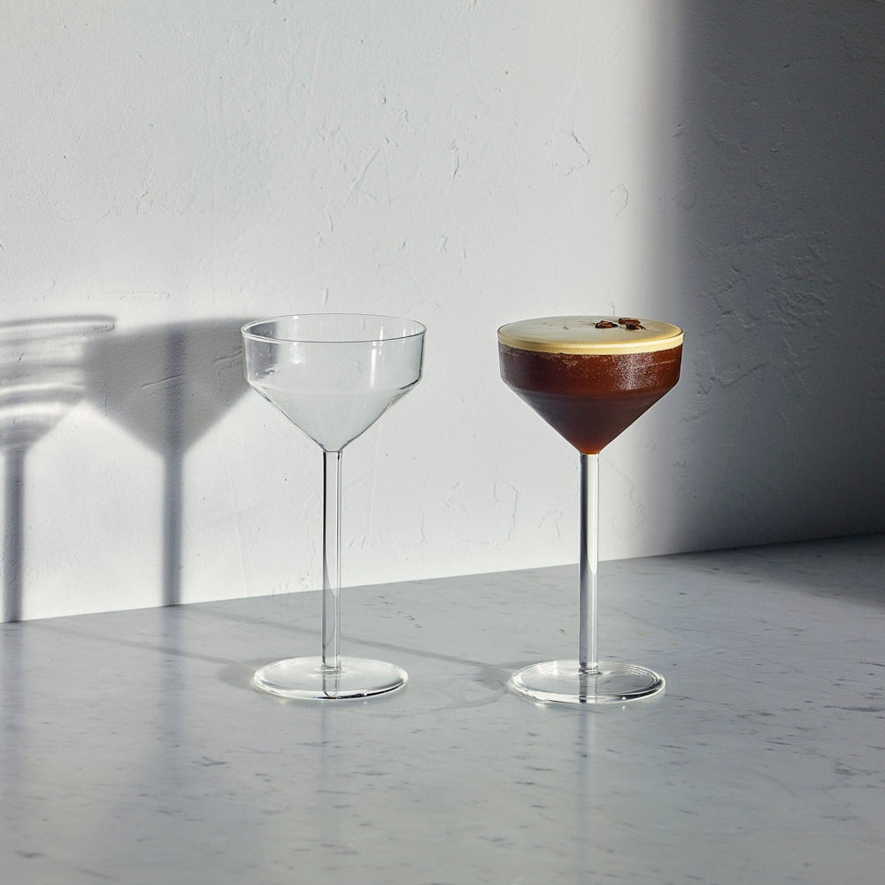 Two clear stemmed cocktail glasses on a light gray surface against a white wall, with one glass empty and the other filled with a dark brown drink topped with a pale foam.
