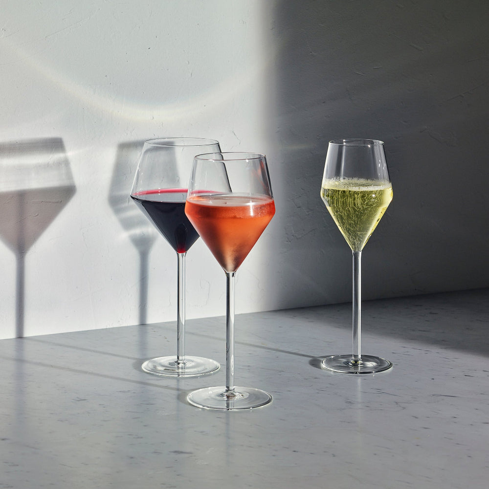 Three stemmed wine glasses with wide bowls are arranged on a gray surface against a gray wall. The left glass contains a dark red liquid, the center glass contains a pink-orange liquid, and the right glass contains a pale yellow bubbly liquid.