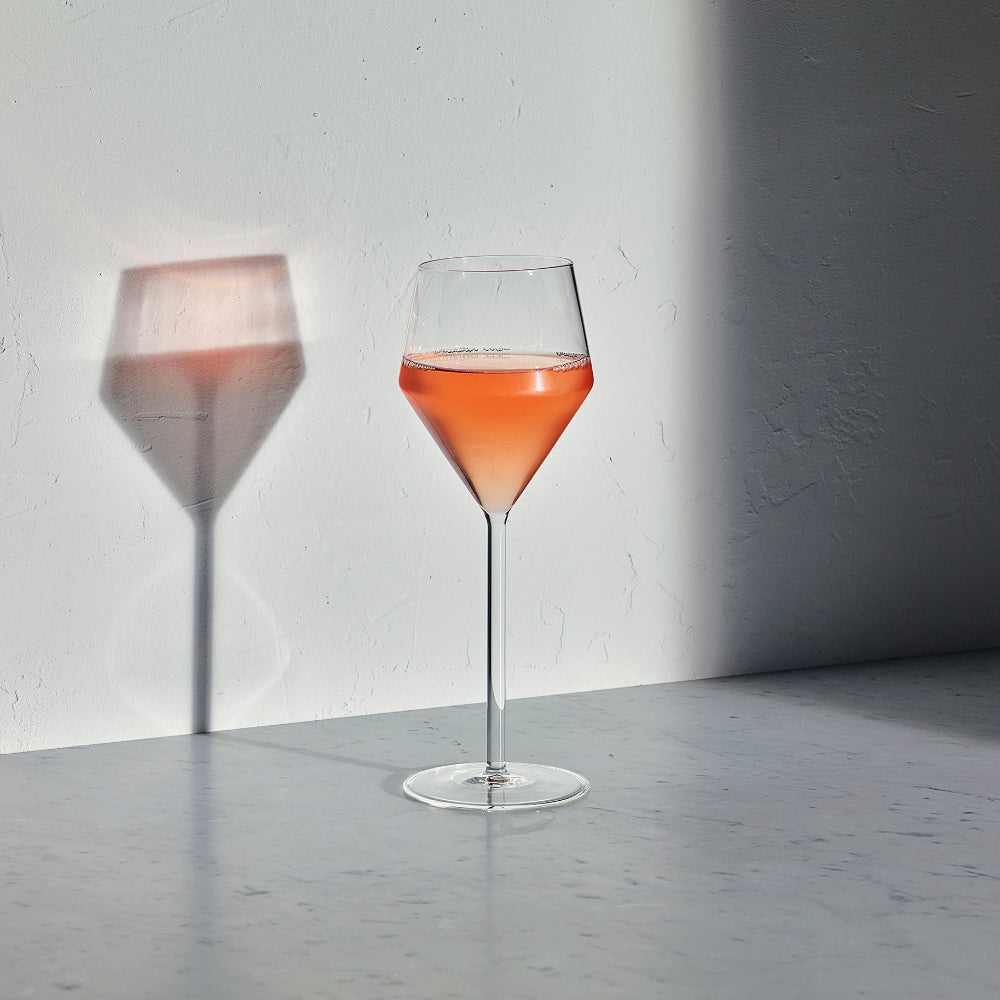 A clear stemmed wine glass with a wide bowl, partially filled with a pink-orange liquid, standing on a light gray surface against a white wall with a strong shadow cast to the left.