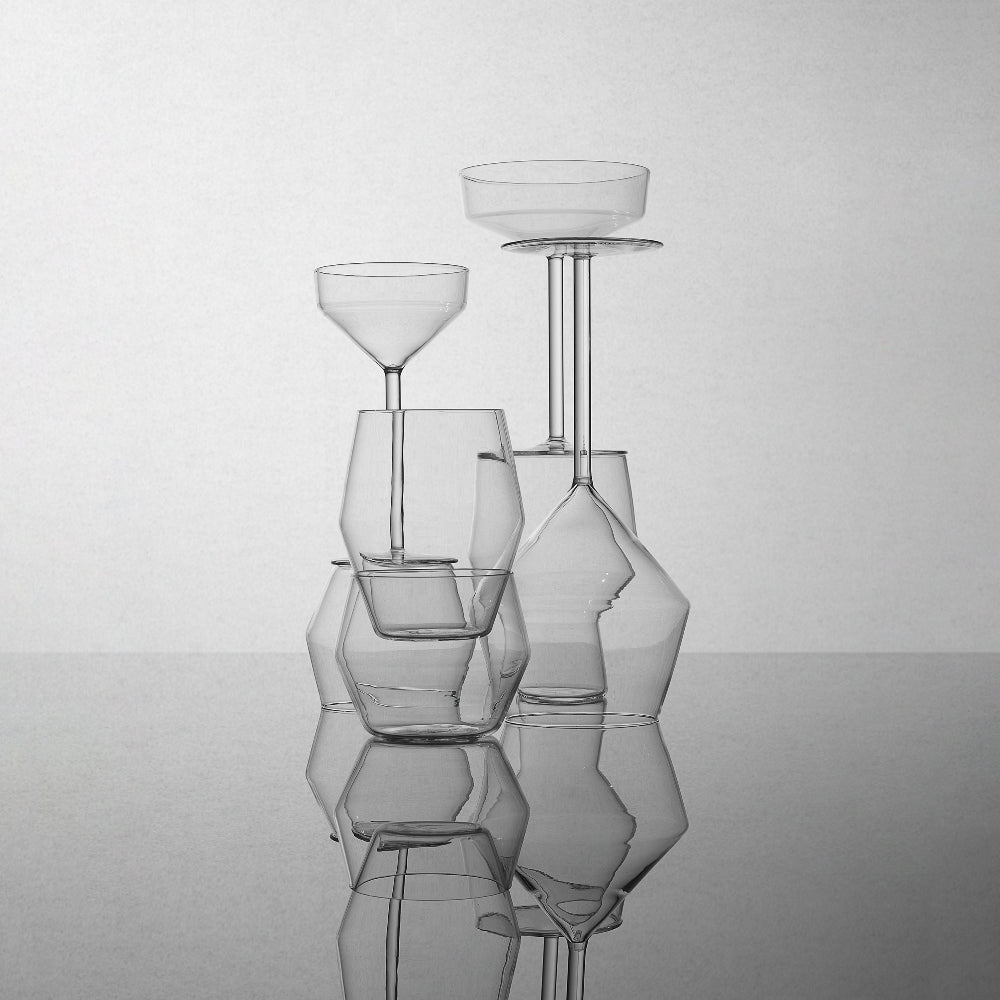 A set of modern, sculptural clear glass drinking glasses arranged on a reflective gray surface against a light gray background.