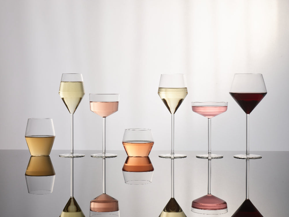 A set of six clear wine and cocktail glasses arranged on a reflective surface against a soft white background, filled with white, pink, and red wines. The glasses include different shapes and sizes, with their reflections visible below.