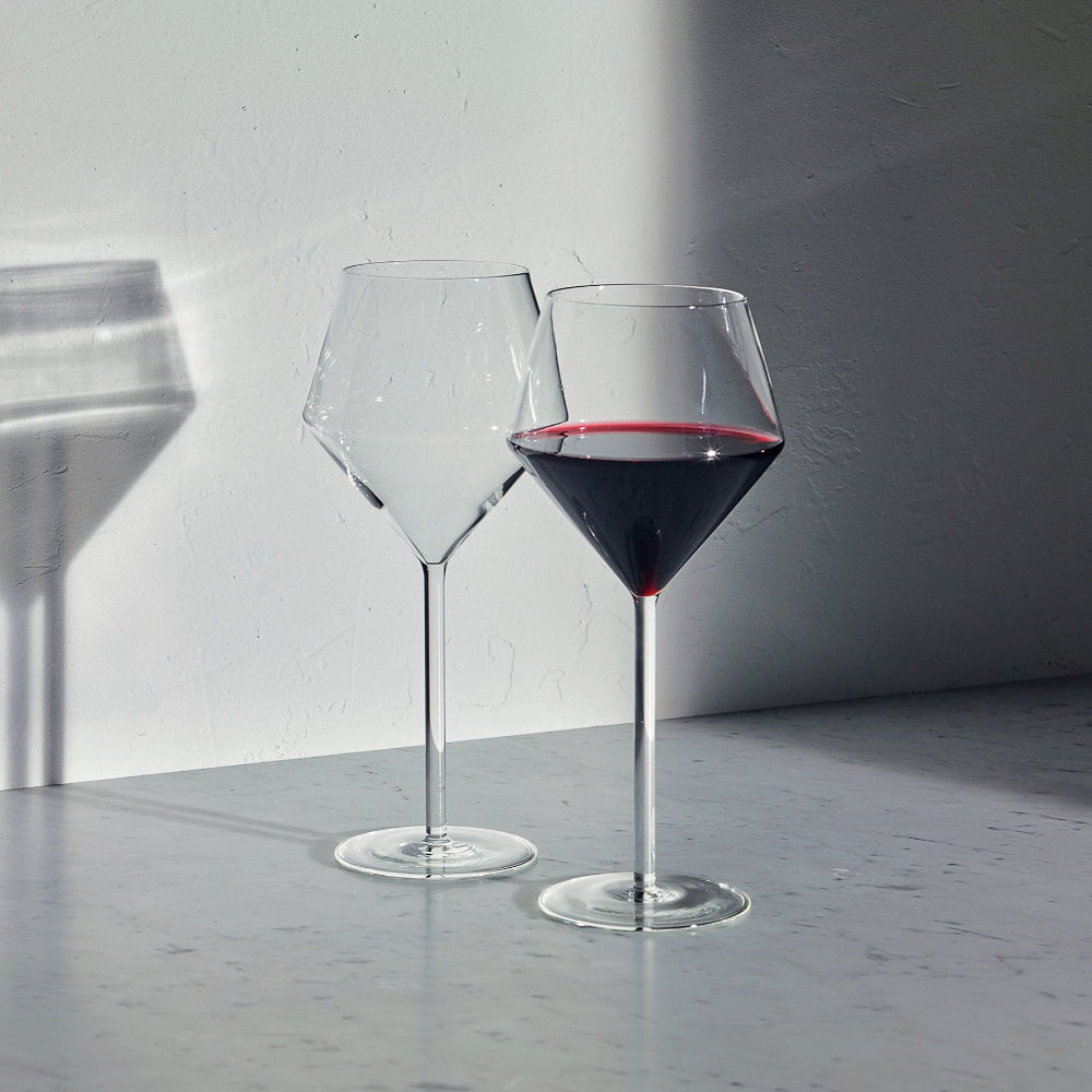 Two clear wine glasses on a gray tabletop against a white wall, with one glass empty and the other partially filled with red wine. Strong sunlight casts sharp shadows across the wall and surface.