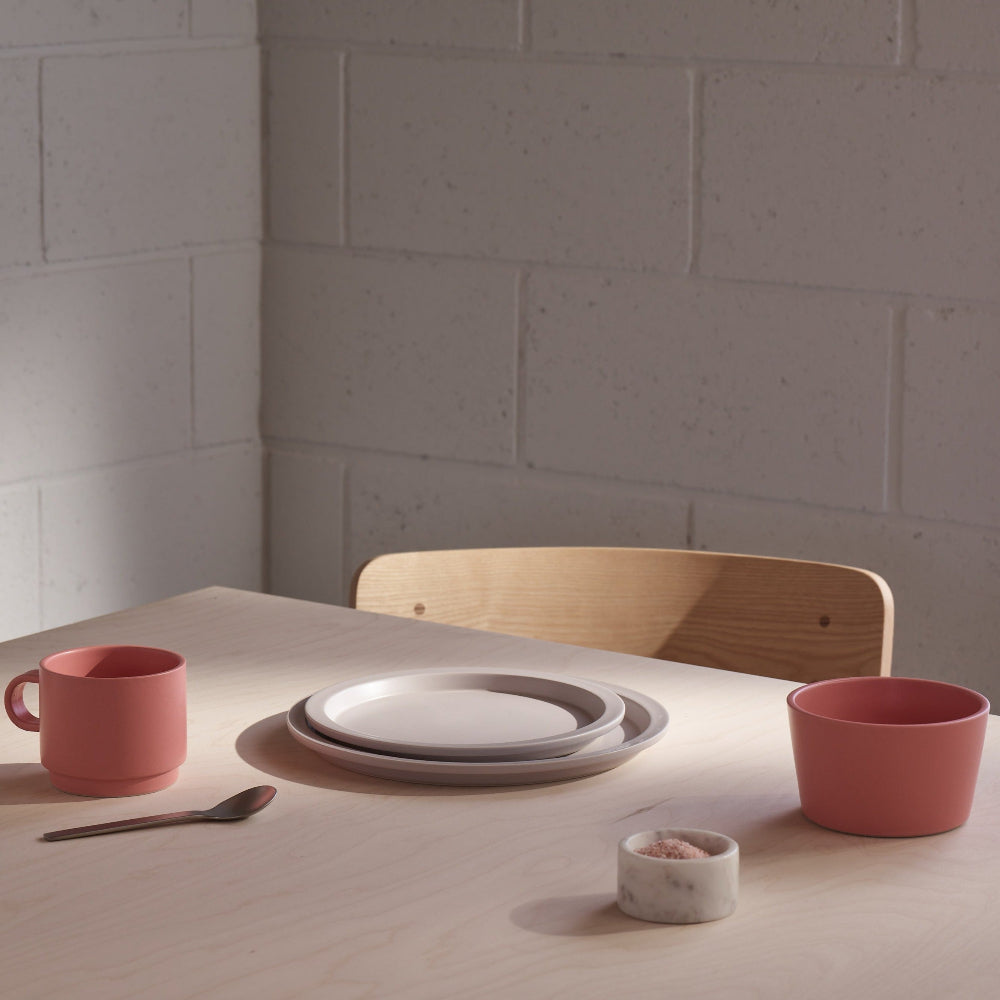 A minimalist dining table setup featuring a dusty pink ceramic mug, matching bowl, white plates, a small marble salt dish, and a wooden chair against a light gray brick wall.