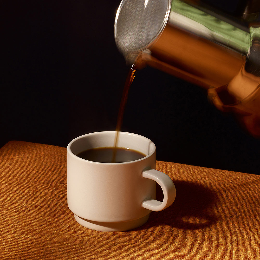 A white ceramic coffee mug sits on a brown surface while coffee is being poured from a metallic pot into it against a dark background.