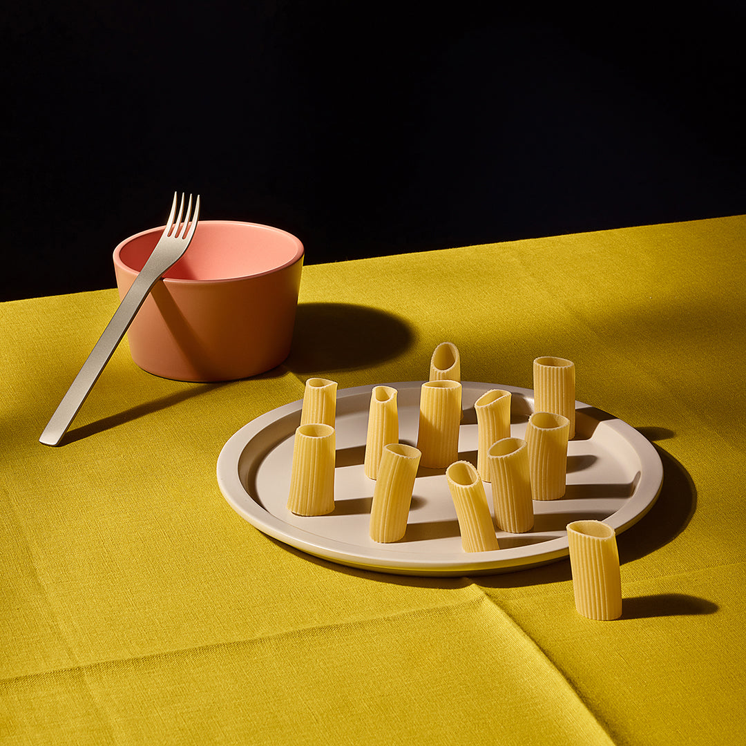 Pasta shapes on a plate with a pink bowl and fork on a yellow tablecloth.