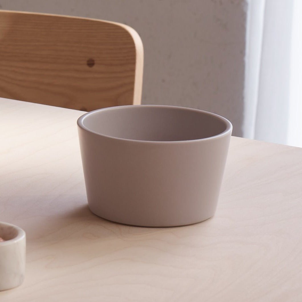 A matte taupe ceramic bowl sitting on a light wood table in a minimalist interior setting.