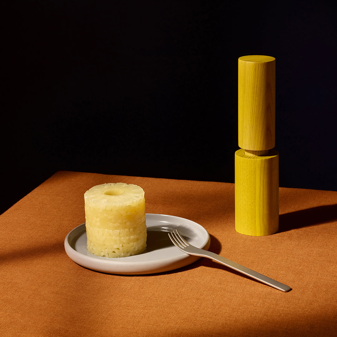 Pineapples sit on a white plate with a fork, next to a matching yellow salt and pepper grinder, on a warm brown surface against a black background.
