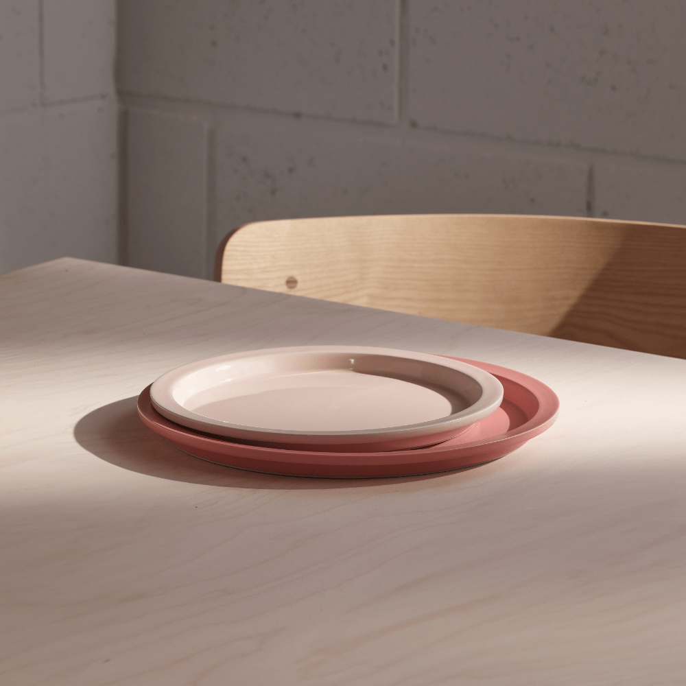 Two nested ceramic plates, one in a matte dusty rose pink and the other in a warm cream, resting on a light wood table in natural sunlight.