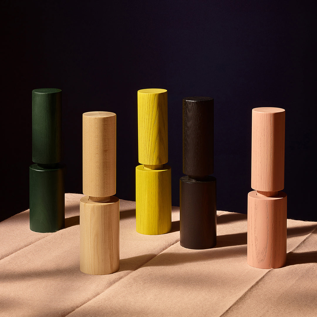 Six wooden cylinder salt and pepper mills in assorted natural and painted colours, stacked in pairs on a beige fabric surface against a dark navy background.
