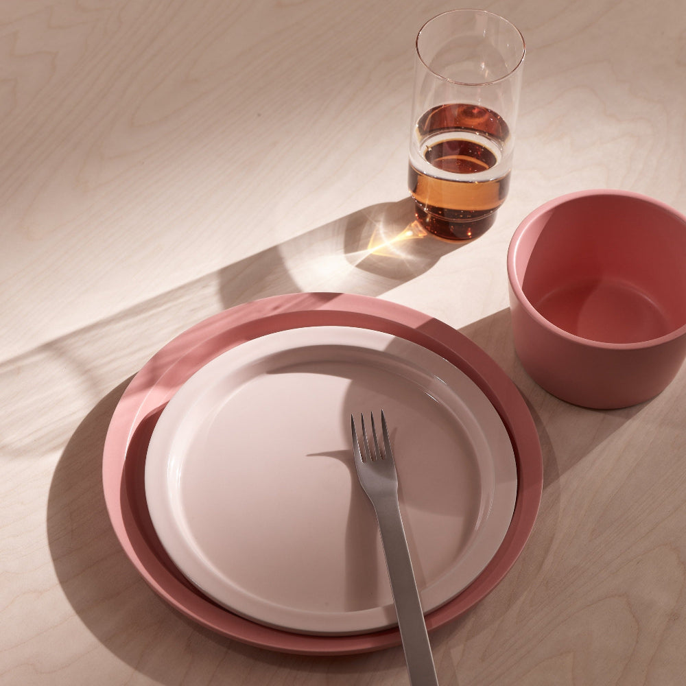 A pink and white ceramic table setting on a light wood table, featuring a stack of plates, a silver fork, a pink cup, and a glass of amber-colored liquid in the background.