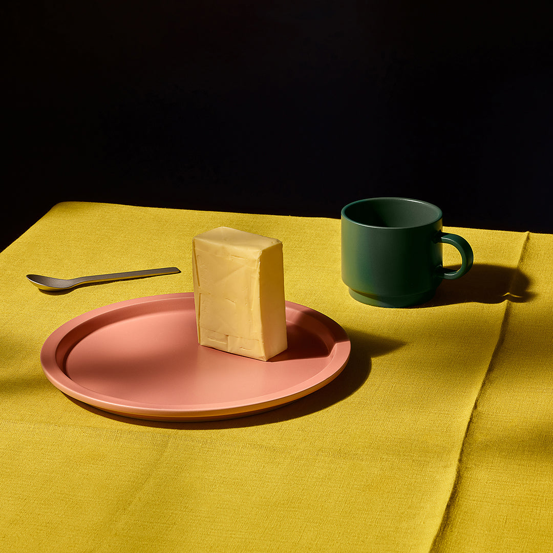 A styled tabletop scene with a pink plate holding a rectangular cream-colored block, a dark green ceramic mug, and a small spoon on a yellow cloth against a black background.