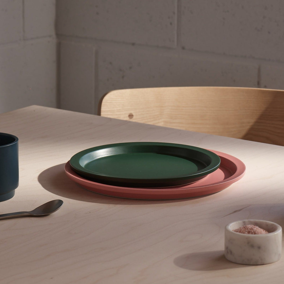 Two matte ceramic plates, one dark green and one dusty pink, stacked on a light wood table in a sunlit dining setting.