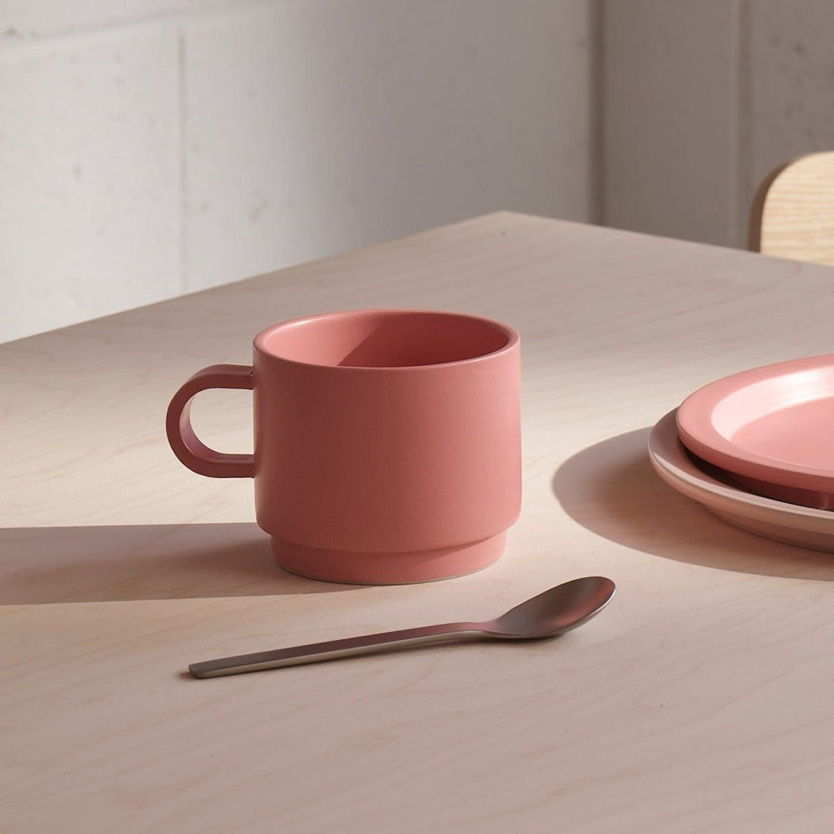 Pink ceramic mug, spoon, and plates on a wooden table with a neutral background