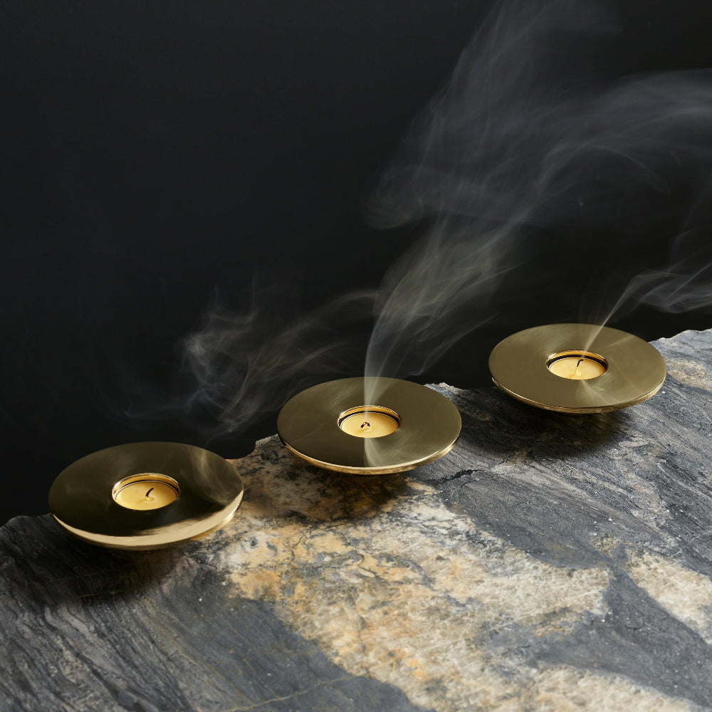 Three round brass candle holders with visible wicks and rising smoke, arranged on a dark stone surface against a black background.