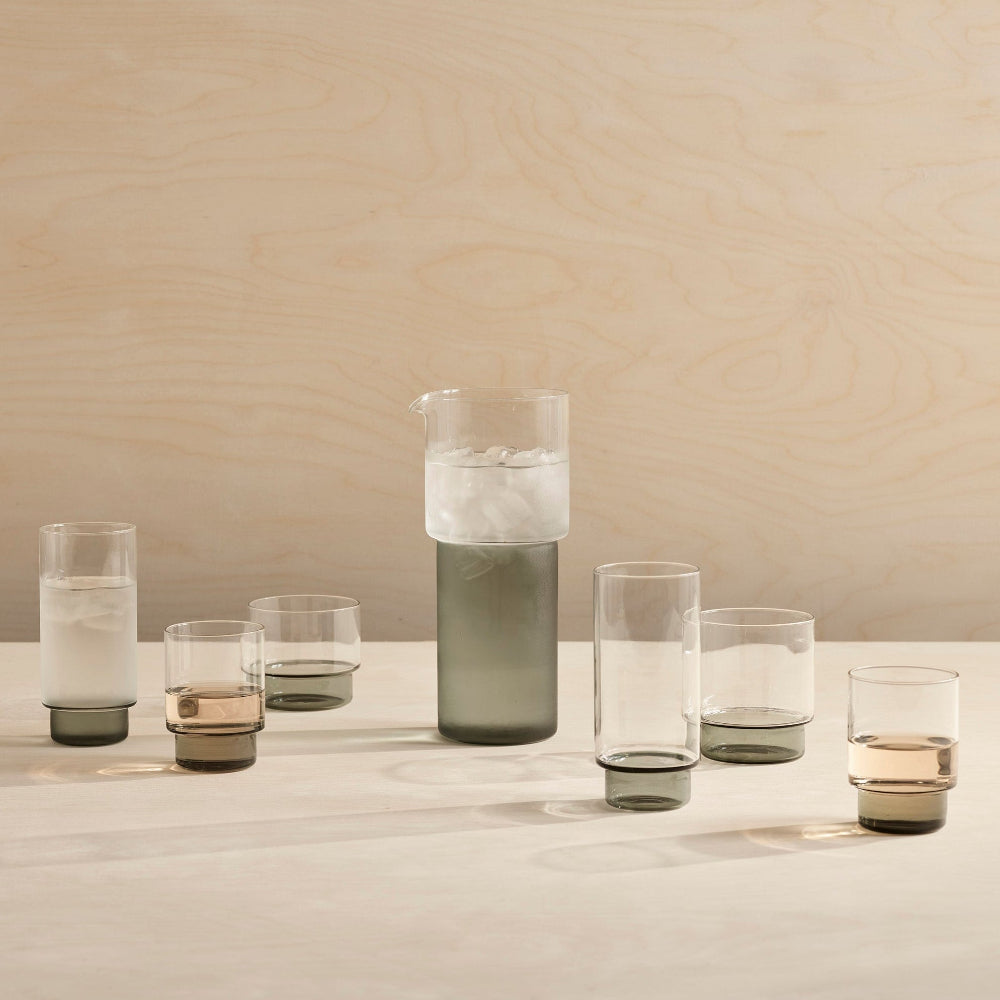 A set of modern smoked glass drinkingware, including a tall carafe with ice and five tumblers of varying heights, arranged on a light wood surface against a matching wood-paneled background.