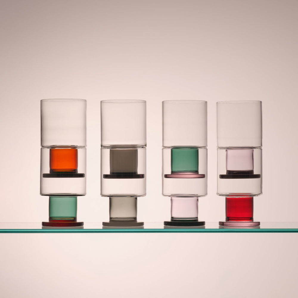 Four modern stacked drinking glasses with clear glass bodies and colorful translucent inserts, arranged on a glass shelf against a warm beige background.