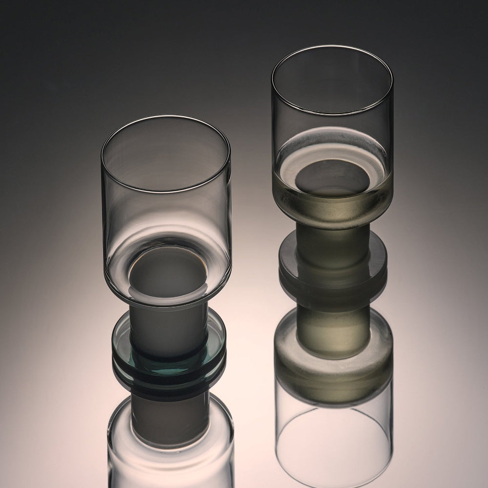 Two modern transparent glass vessels with stacked cylindrical forms and reflective surfaces, photographed on a smooth dark-to-light gray gradient background.