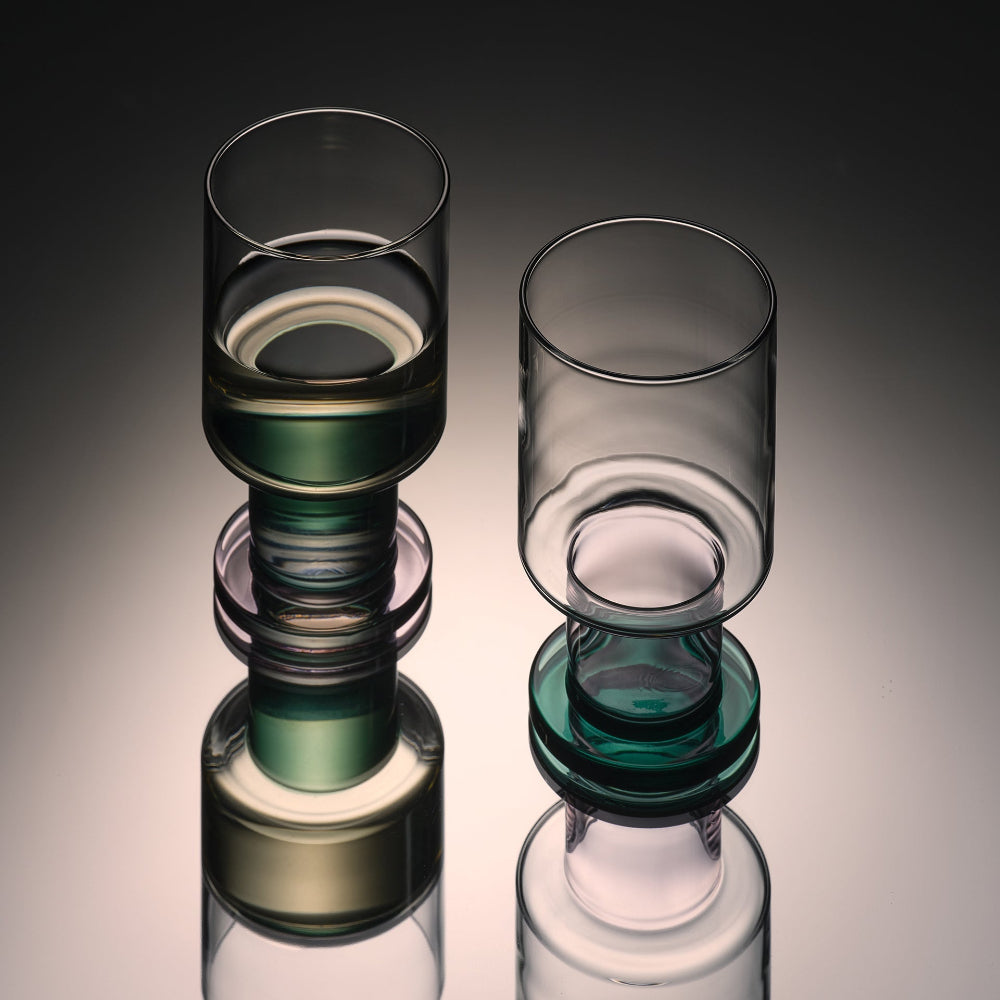 Two modern stacked glass tumblers with translucent dark green and clear glass cylindrical sections, displayed on a reflective surface against a dark gray gradient background.