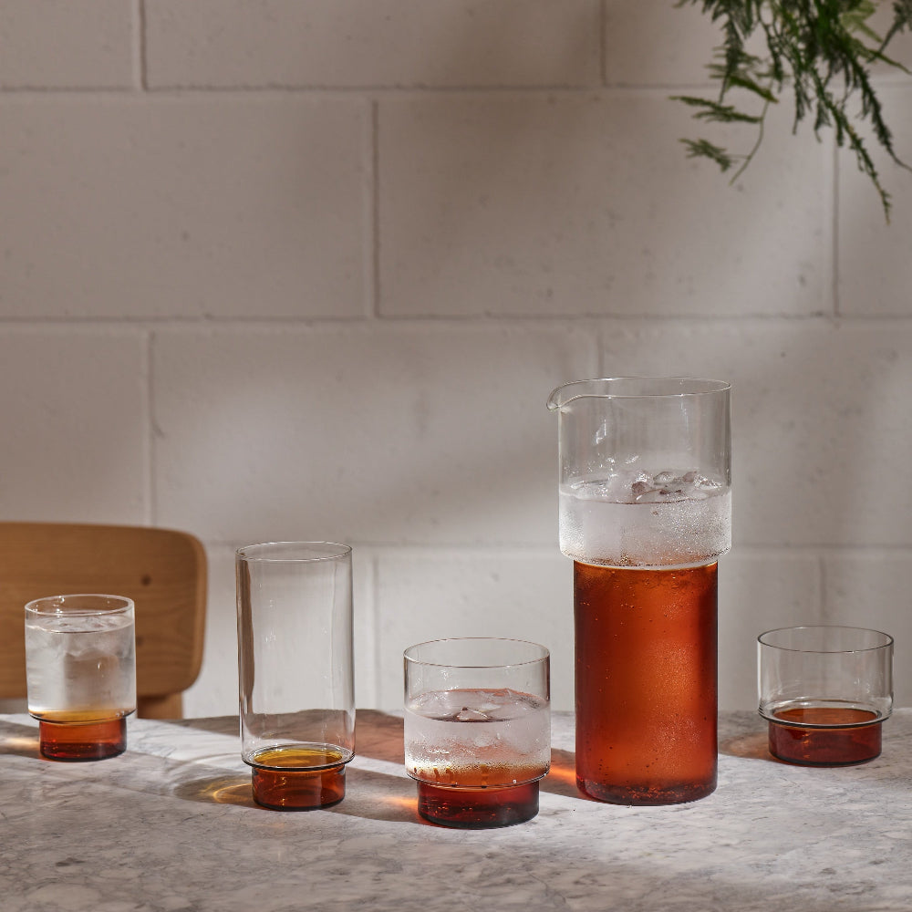 A set of clear glass drinking glasses and a matching glass carafe with amber-colored liquids and ice, arranged on a stone surface against a light brick wall.