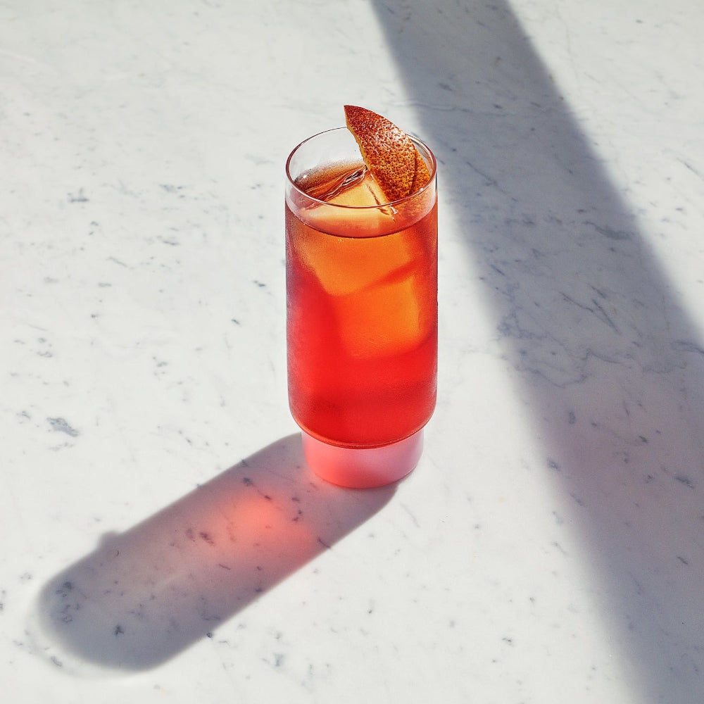 A tall clear glass filled with a bright red-orange drink and a citrus garnish, set on a white marble surface with strong sunlight and shadows.