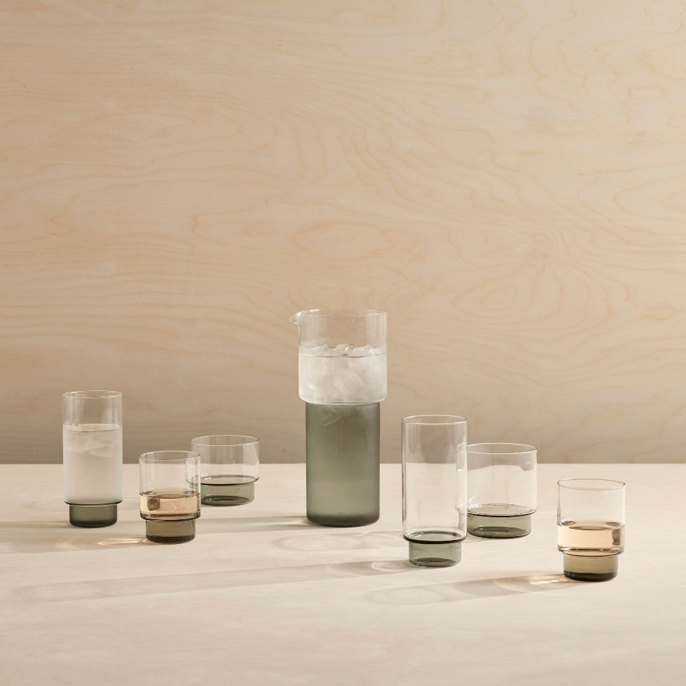 A set of modern glassware including a tall water carafe and five tumblers of varying heights and widths, featuring smoky translucent dark gray and amber-brown glass accents, arranged on a light wood surface against a matching wood-paneled background.